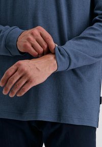 Langarmhemd aus strukturierter blauer Stoff. Hat eine lockere Passform, gerippte Säume und einen geraden Saum. Hände, die den Ärmel anpassen, sind sichtbar.