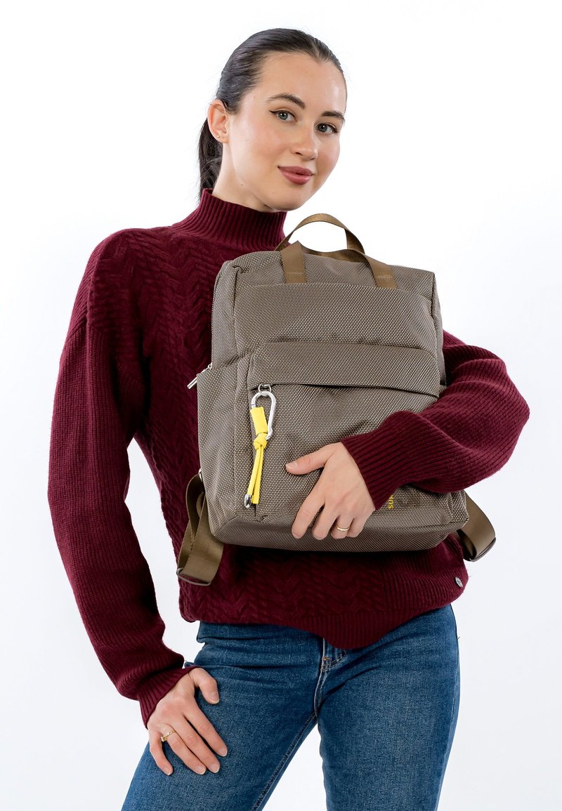 Ruskea teksturoitu reppu, jossa on kaksi yläkahvaa, säädettävät hihnat ja keltainen vetoketjun vedin. Kuvattuna valkoisen taustan edessä.