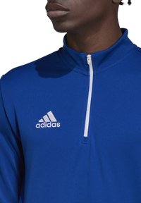 Blauwe, ritssluiting sportjack gemaakt van een gladde stof, met een hoge kraag en het witte Adidas-logo op de linkerborst.