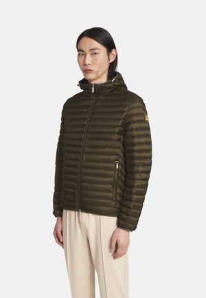 Man draagt een olijfgroene doorgestikte donzen jas met kap en beige broek, staand tegen een effen lichte achtergrond.