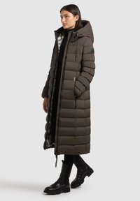 Giacca imbottita lunga marrone con cappuccio, rivestimento interno argentato lucido, tasche con zip e design trapuntato. Indossata con stivali neri.