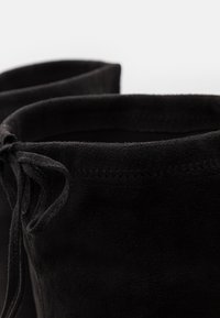 Stivaletti neri in suede con una texture morbida, caratterizzati da un bordo superiore arrotondato e un dettaglio di lacci su un lato. Cucitura lungo l'apertura.