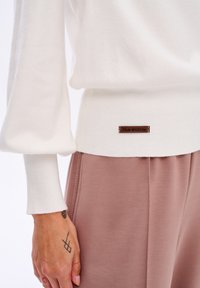 Biały sweter z długimi, bufiastymi rękawami, ściągaczami przy rękawach i u dołu oraz brązową metką w okolicy talii; zestawiony z jasnoróżowymi spodniami.