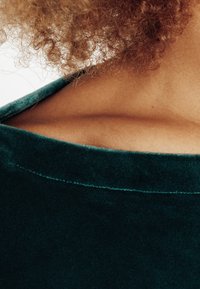 Tessuto di velluto verde con una consistenza morbida, che mostra parte di un colletto arrotondato e pelle. Dettagli di cucitura vicino al colletto.