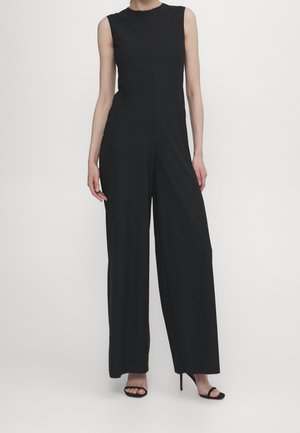 Kvinde iført en ærmeløs sort jumpsuit med vide ben og høj hals, samt sorte åbne højhælede sandaler, stående mod en ensfarvet baggrund.