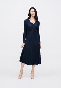 Tengerészkék hosszú ujjú ruha csavart elülső dizájnnal, pliszírozott textúrával és középmély szoknyával; világos színű, hegyes orrú magassarkúval párosítva.