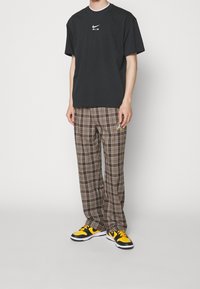 Svart bomullst-shirt med vit Nike-logga, tillsammans med bruna rutiga byxor med en sidodekor och svarta sneakers med gula accenter.