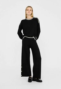 Zwarte gebreide trui met witte afwerking en een ontspannen pasvorm, gecombineerd met zwart wijde broekspijpen met knoopaccenten aan de zomen.