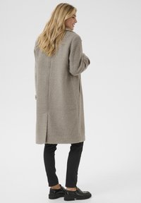 Vezelige, lichtgrijze jas met een comfortabele pasvorm en zijzakken, gedragen over zwarte slim-fit broeken en stevige zwarte instappers. Achteraanzicht weergegeven.