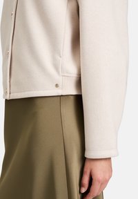 Jasnobeżowa kurtka z miękkiego, teksturowanego materiału, z zapięciem na guziki i ściągaczem u dołu, w połączeniu z gładką oliwkowozieloną spódnicą.