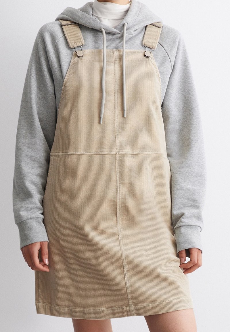 Beige kordfluga overallklänning med justerbara axelband, kombinerad med en grå hoodie. Har vertikal ribbning och en rak passform.