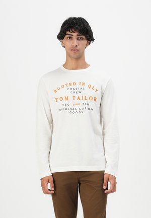 Junger Mann mit dunklem, lockigem Haar, der ein weißes Langarmhemd mit orangefarbener und schwarzer Schrift sowie braunen Hosen trägt und vor einem weißen Hintergrund steht.