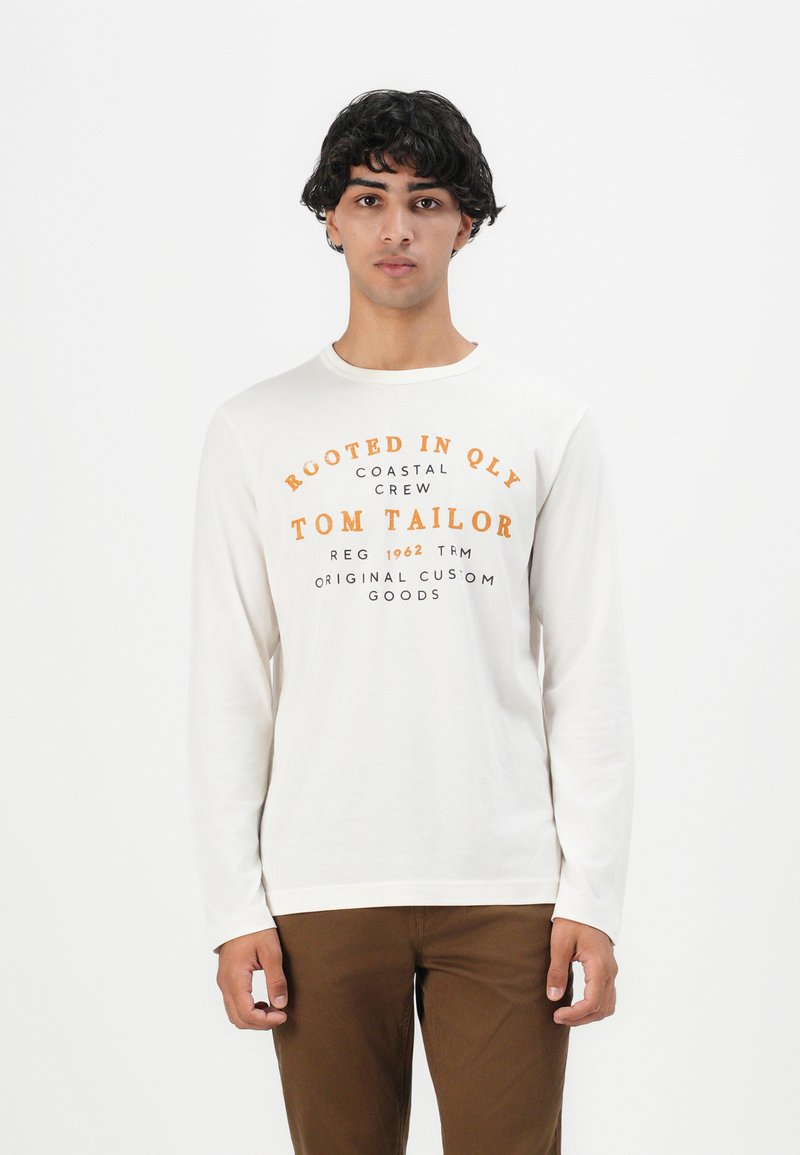 Νεαρός άνδρας με σκούρα σγουρά μαλλιά, φορώντας ένα λευκό μακρυμάνικο πουκάμισο με πορτοκαλί και μαύρο κείμενο και καφέ παντελόνι, στέκεται μπροστά σε λευκό φόντο.