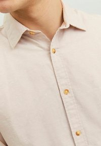 Vaaleanpunaista napitettua paitaa, jonka kauluksessa on kaulus ja kultaiset nappikoristelut etupuolella, valmistettu teksturoidusta kankaasta.