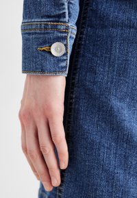 Μανίκι από τζιν μπουφάν με ασημένιο κουμπί, χρυσές ραφές και λεία υφή. Το μπλε ύφασμα παρουσιάζει ένα τυπικό σχέδιο τζιν.