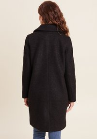 Zwarte wollen overjas met een hoge kraag, lange mouwen en een rechte snit. Kenmerkt zich door een gestructureerde stof en een licht gebogen zoom aan de achterkant.