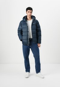 Mørkeblå pufferjakke med hette, med frontglidelås, quiltet tekstur og logo på brystet. Kombinert med lyseblå jeans og hvite sko.