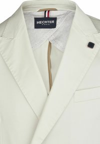 Herren Creme-Blazer mit spitzen Revers, einem gemusterten Innenfutter, einer kleinen schwarzen Akzentnadel und einer einzelnen Brusttasche.