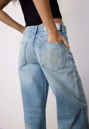 Kobieta ubrana w jasnoniebieskie dżinsy z kryształowymi gwiazdkami na tylnych kieszeniach, ręka w prawej kieszeni, widoczny krótki czarny top.