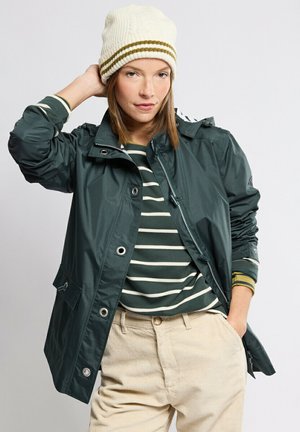 Femme portant un bonnet en tricot blanc, une chemise à rayures vertes, une veste vert foncé et un pantalon beige, une main derrière la tête et l’autre dans la poche.