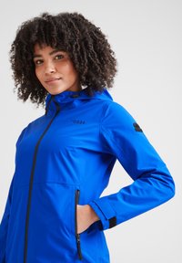 Woman with curly hair wearing a bright blue TOG24 hooded jacket, hand in zipped pocket, facing slightly to the right with a neutral background.