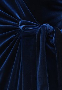 Zoom na temno modro žametno tkanino, zbrano in zvezano v mehak vozel z gladkimi gubami in bogato teksturo.