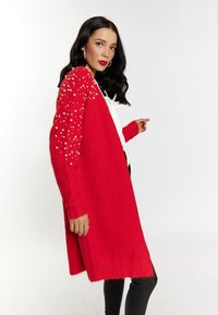 Cardigan rosso lavorato a maglia con decorazioni in perle sulle maniche, lunghezza lunga e polsini a coste, abbinato a una maglietta bianca e pantaloni neri slim.