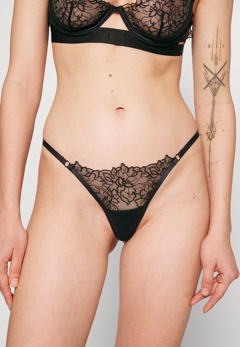 Fio-dental preto em renda com bordados florais na parte frontal, painéis em malha, alças finas e pequenos detalhes em anéis. Textura suave e cobertura mínima.