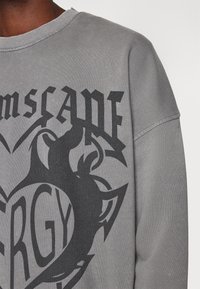 Sudadera gris hecha de una mezcla de algodón. Presenta un gráfico en negro audaz con texto y elementos de diseño, incluidos formas curvadas y contornos.