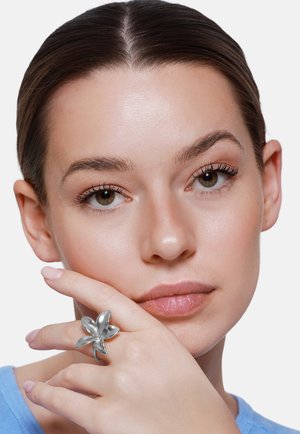 Jeune femme à la peau lisse et aux yeux marron, reposant son menton sur sa main, portant une grande bague en argent en forme de fleur.