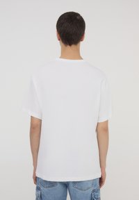 Biała bawełniana koszulka z krótkim rękawem i okrągłym dekoltem, pokazana z tyłu, charakteryzująca się luźnym krojem i prostym wykończeniem.