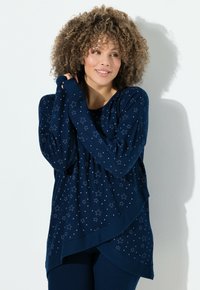 Frau mit lockigem Haar trägt einen navyblauen Pullover mit Sternmuster und passende Hose, lächelt und hält die Pulloverärmel nah an ihr Gesicht.