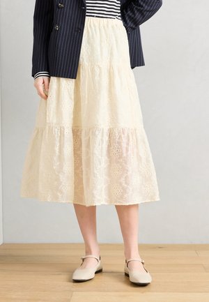 Kvinne iført kremfarget midiskjørt i blonder, marineblå blazer med tynne striper, stripet topp og beige Mary Jane-sko, stående på tregulv.