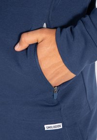 Felpa blu navy dal design aderente. Presenta una tasca con zip sul lato e una piccola etichetta bianca del marchio sull'orlo. Tessuto morbido.