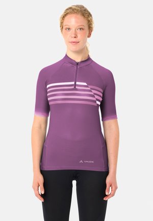 Frau trägt ein lila kurzärmeliges Radtrikot mit horizontalen Streifen und schwarze Radhosen, steht vor einem weißen Hintergrund.