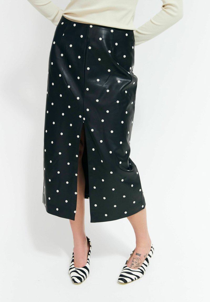 Jupe midi noire à pois blancs avec une fente sur le devant, portée avec des talons hauts à rayures zébrées et un haut à manches longues de couleur claire.
