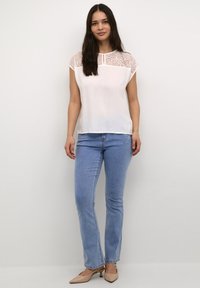 Blusa bianca con sovrapposizione in pizzo e maniche corte, abbinata a jeans a gamba dritta azzurri chiari e tacchi bassi beige, in piedi di fronte a uno sfondo semplice.
