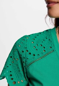 Top verde con mangas de encaje detalladas y patrones recortados, con un cuerpo sólido. Diseño de escote en V, textura suave en toda la prenda.