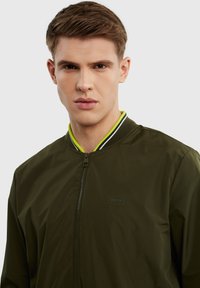Alkšninė bomber tipo striukė su užtrauktuku, turinti juodą ir neoninės žalios spalvos raištelį, bei mažą logotipą krūtinėje.