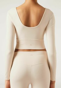 Beige Langarm-Crop-Top mit weit ausgeschnittenem Rücken und nahtlosem Design, kombiniert mit passenden hochtaillierten Leggings mit dezentem Branding.
