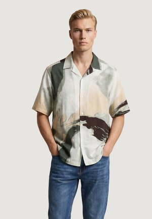 Uomo biondo in piedi con le mani nelle tasche, indossa una camicia a maniche corte con bottoni con motivi astratti beige, verdi e marroni e jeans blu.