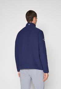 Sweater azul-marinho com gola alta, textura canelada e detalhe de logótipo na parte superior do braço. Tecido em mistura de algodão com corte descontraído.