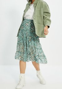 Jupe florale vert clair avec un design en étages, fabriquée dans un tissu transparent. Assortie à une veste verte surdimensionnée et des bottes blanches à lacets.