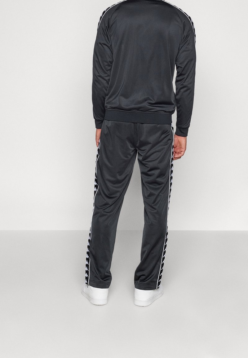 Homme portant un survêtement noir avec des bandes blanches de logo le long des manches et des jambes du pantalon, debout de dos sur un sol gris et un fond blanc.
