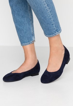 Personne portant un jean bleu court et des ballerines en daim bleu foncé, debout sur un sol gris clair.