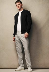 Černá pletená bomber bunda přes bílé tričko, spárovaná s světle šedými šitými kalhotami a bílými teniskami. Minimalistický design, pohodlný střih.