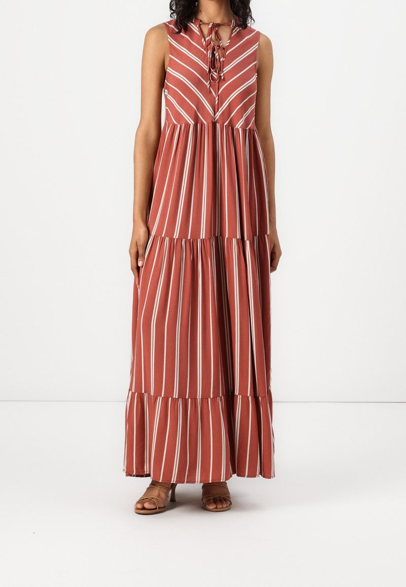 Robe maxi sans manches de couleur rouille avec des rayures verticales blanches, jupe à volants et col à nouer, portée avec des talons ouverts marron.