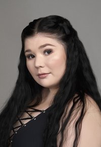 Portrait d'une personne avec de longs cheveux noirs ondulés. Ils portent un haut noir avec des bretelles croisées et des accents métalliques. Fond neutre.