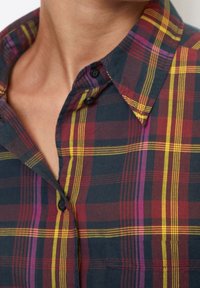 Camicia a quadri con una base scura e linee rosse, gialle e viola. Il colletto con bottoni ha una forma strutturata e un tessuto testurizzato.