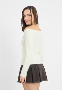 Cremfarbener, schulterfreier Pullover aus flauschigem Material, kombiniert mit einem dunklen, plissierten Minirock mit fransigem Saum.